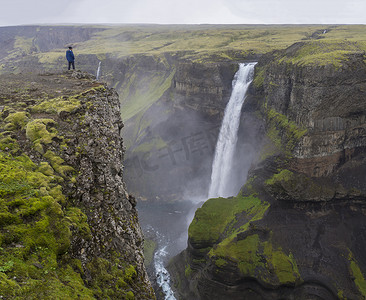 佛身摄影照片_冰岛南部的 Fossa 河谷和美丽的 Haifoss 瀑布，身穿蓝色夹克的男子站在悬崖岩石上看风景