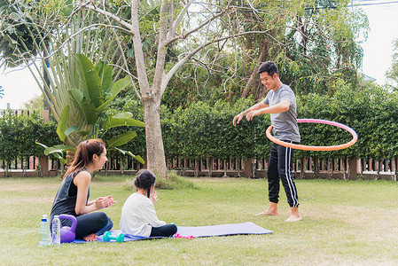 孩子加油摄影照片_母亲和孩子的女儿在户外练习瑜伽练习为玩呼啦圈的父亲加油