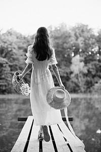 一位身穿浅色长裙的女子背着篮子，头戴柳条帽，站在森林湖边的码头上。