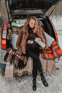金发女人裹着毯子坐在后备箱里喝着热水瓶里的咖啡茶。