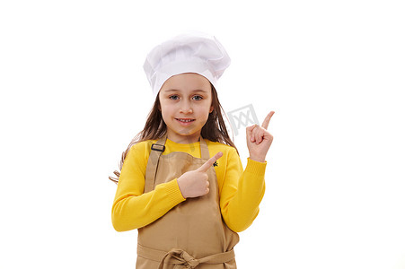 戴着厨师帽和米色厨房围裙的微笑快乐的小女孩，用手指指着白色背景中的复制广告空间