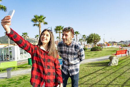 年轻快乐的情侣在日落或黎明时分在海洋度假村的城市公园里用智能手机微笑自拍肖像。