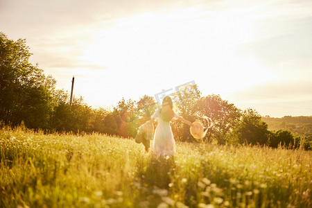温暖亲情摄影照片_一位穿着浅色连衣裙的女士站在田野里，沐浴在夕阳的温暖光线中，头上戴着一顶帽子