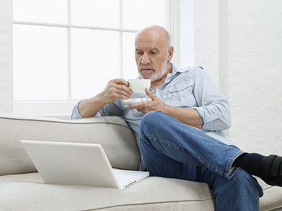 边顶摄影照片_微笑的成熟男人边喝咖啡边在家沙发上看笔记本电脑
