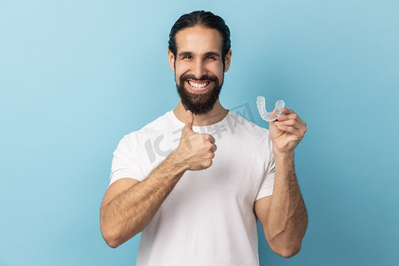 固定假牙摄影照片_男人拿着牙齿矫正器固定器，美丽牙齿的牙科诊所，竖起大拇指。
