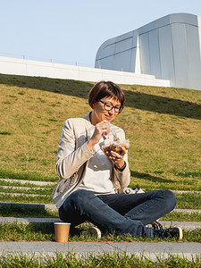 “女人坐在公园长椅上，拿着带走的午餐盒和纸杯”