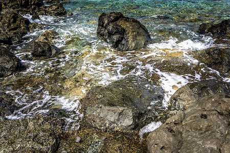 清澈蓝色水摄影照片_黑色的石头和清澈的海水在海滩上冲浪。