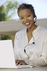 一位微笑的年轻支持电话接线员的肖像，他戴着耳机，在露台上带着笔记本电脑