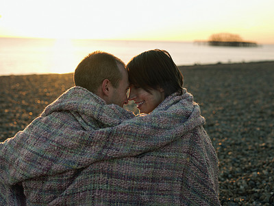 拥抱恩爱摄影照片_日落时分，一对恩爱的情侣在海滩上裹着毯子的侧视图