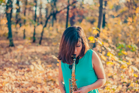 身穿蓝色裙子的黑发少女在黄秋林中热情地演奏萨克斯管