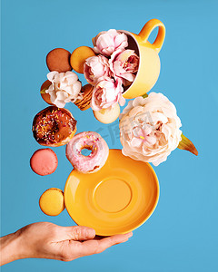 黄色杯子里的牡丹、甜甜圈、马卡龙、牡丹和蓝色背景上飞舞的手托碟子的静物画
