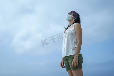 冠状病毒海滨假期：一名妇女在海滩上的照片，她戴着面具看着太阳，因为 Covid-19 大流行病和多云的天空