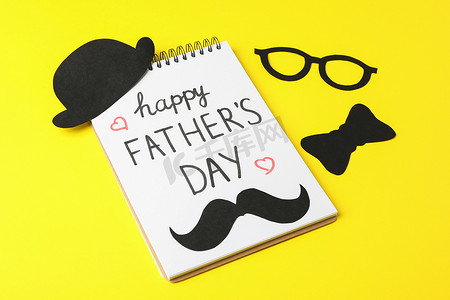 父亲节文字背景摄影照片_笔记本上刻有父亲节快乐、装饰性领结、眼镜、胡须和彩色背景上的帽子、文字空间