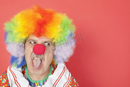 小丑背景摄影照片_在红色背景中望向别处时伸出舌头的高级男性小丑