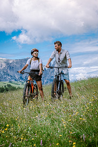 情侣在意大利多洛米蒂山度假，Alpe di Siusi，男人和女人骑 mountanbike - Seiser Alm 与 Sassolungo - Langkofel 山群在日落时的背景。