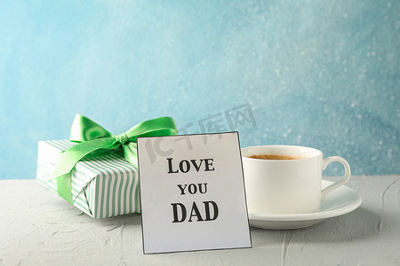 一杯咖啡、带绿丝带的礼盒和题词“爱你爸爸”在蓝色背景的白桌上，文字空间