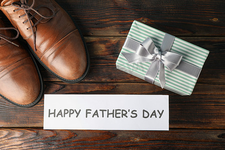 父亲节文字背景摄影照片_棕色皮鞋、快乐父亲节的铭文和木制背景上的礼盒、文字空间和顶视图