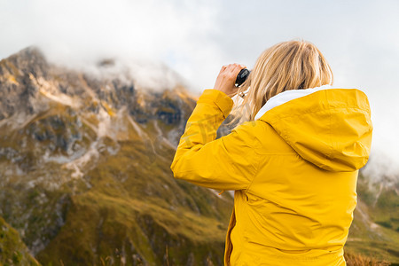 年轻的金发女子在山中徒步旅行并通过双筒望远镜观察