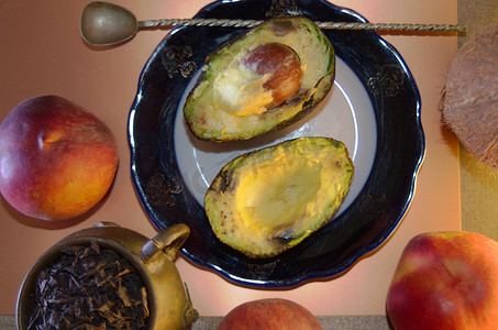 食物成分——盘子上的两半鳄梨和一把扭曲的勺子、三个成熟的桃子、一个装有大叶茶和椰子的古董铜杯