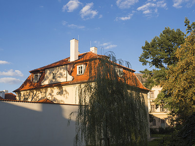 金色城市背景摄影照片_Kampa 的布拉格巴洛克式老房子有红色屋顶和白色围墙，黄金时段的开窗，树木和蓝天背景