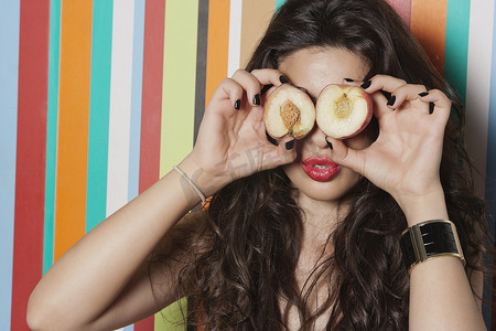 眼睛横截面摄影照片_条纹背景下用桃子遮住眼睛的年轻女人