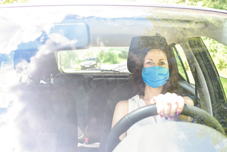 防护汽车摄影照片_一位戴着防护手术面具的成年女司机坐在车里，看着相机