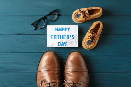 父亲节文字背景摄影照片_棕色皮鞋、童鞋、父亲节快乐的铭文、木质背景的眼镜、文字空间和顶视图