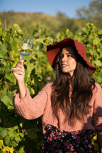 看葡萄酒的颜色摄影照片_戴着时髦帽子的年轻女性看着酒杯里葡萄酒的颜色