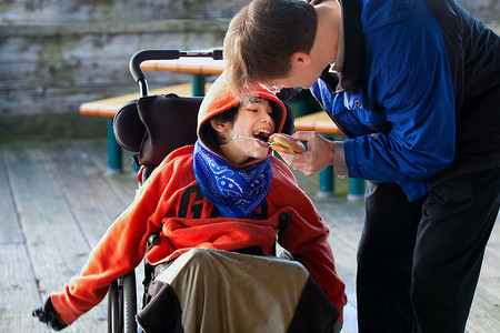 儿子给父亲摄影照片_父亲给坐在轮椅上的残疾儿子喂汉堡包。
