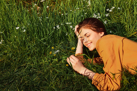 一个长着红色长发的平静女人躺在开着黄色花朵的绿色田野里，身穿橙色连衣裙，愉快地微笑着，在灿烂的夏日阳光下闭上眼睛，将头靠在双手上