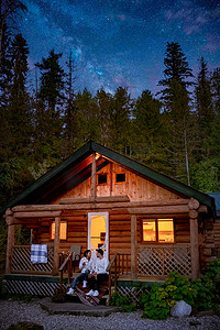 加拿大不列颠哥伦比亚晚上在树林里的小屋，情侣在木屋前喝酒，银河前的偏远小屋
