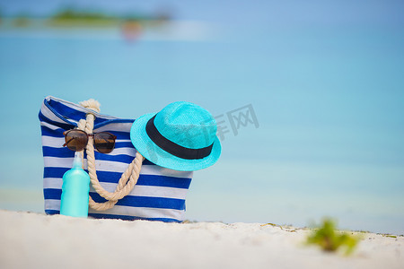 白色沙滩上的蓝色包、草帽、太阳镜和防晒瓶