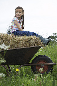 坐在推车的女孩摄影照片_女孩（5-6 岁）坐在手推车的干草上，在田野肖像中