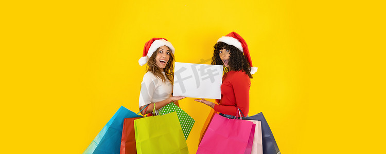 “两个戴着圣诞老人帽的年轻可爱女人面带惊讶地看着镜头，张开嘴，睁大眼睛，拿着彩色购物袋-带有黄色背景的圣诞横幅，用于复制空间”