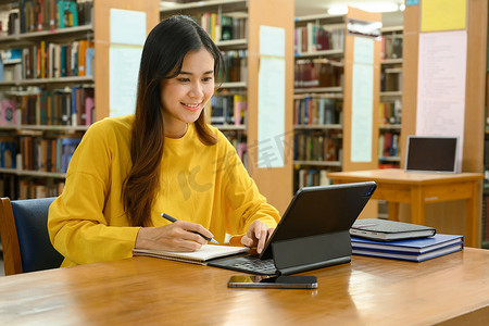 快乐的亚洲女学生参加在线课程，在图书馆用数字平板电脑学习。