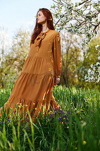 一个苗条、甜美的女人穿着橙色长裙站在一棵开花的树旁高高的草丛中，看着别处，她的裙子在微风中展开