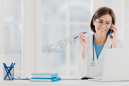 高兴的女治疗师或医生的照片与病人进行电话交谈，提供如何治疗疾病的建议，穿着白色长袍，在笔记本电脑上阅读信息，在私人诊所工作
