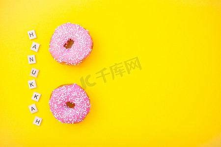 光明摄影照片_黄色背景中的粉红色美味甜甜圈和木块上的铭文光明节快乐