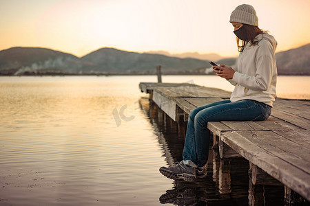 孤独的女人在日落或日出时坐在码头上，用智能手机关注 Covid-19 的最新消息。