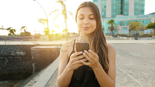 海边女子摄影照片_微笑的年轻女子在日落时分用智能手机发送消息，背景是海边小镇。