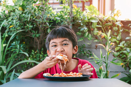 身穿红衬衫的亚洲可爱男孩开心地坐着吃披萨。
