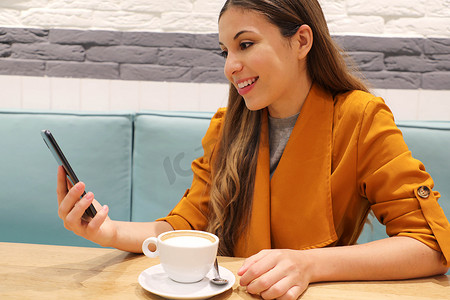 咖啡馆城市生活方式女孩坐在时尚的城市咖啡馆里，在智能手机应用程序上阅读手机喝卡布奇诺的信息