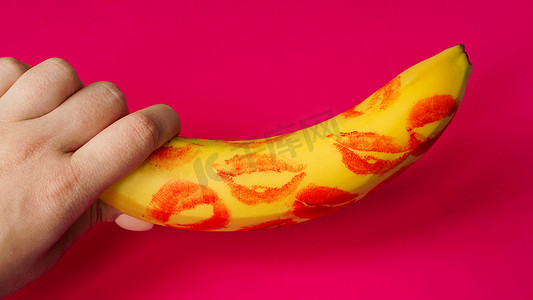 粉红色背景上带有红色唇膏标记的女人手拿着香蕉