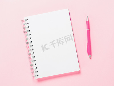 粉色笔记摄影照片_粉红色背景上带笔的空白笔记的顶部视图