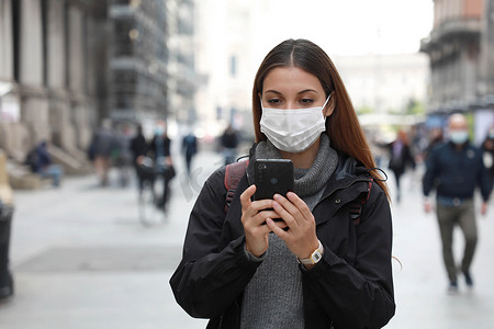 街上戴口罩的人摄影照片_戴外科口罩的漂亮女大学生走在街上用手机