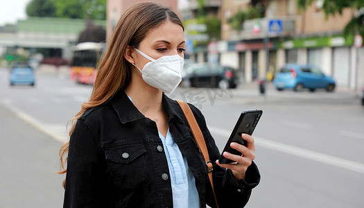 COVID-19 年轻女性佩戴 KN95 FFP2 口罩在城市街道使用智能手机应用软件帮助接触者追踪和自我诊断以应对 2019 年冠状病毒大流行