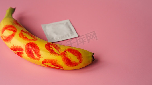 疾病预防背景摄影照片_粉红色工作室背景中黄色香蕉上未包装的避孕套和红色唇膏