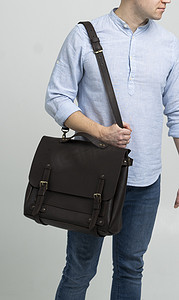 肩包摄影照片_身穿蓝色衬衫和白色背景牛仔裤的男士肩上背着棕色男士肩包，用于存放文件和笔记本电脑。