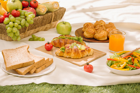 野餐柳条篮，里面有食物、面包、水果和橙汁，放在红色和白色的格子布上，背景是绿色。