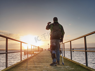 海滩渡口摄影照片_手拿手机的男人在渡口鼹鼠上拍日出照片